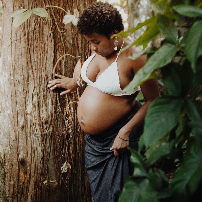 A black pregnant women with her belly bare standing next to a tree.