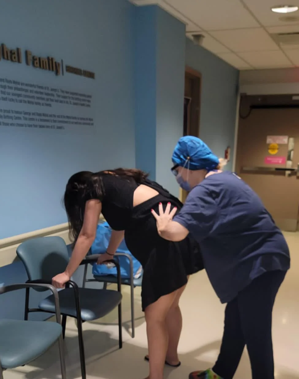 Marcia doing counter pressure during labor. Both mom and doula are standing in a hallway with the mom standing, leaning over a chair and Marcia squeezing her hips together.