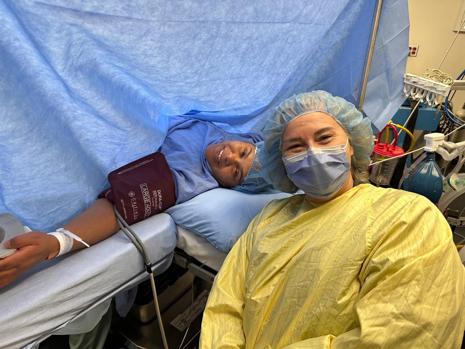 A women and her doula during a c-section. The new momma is lying down draped from her neck down giving birth via cesarean. 