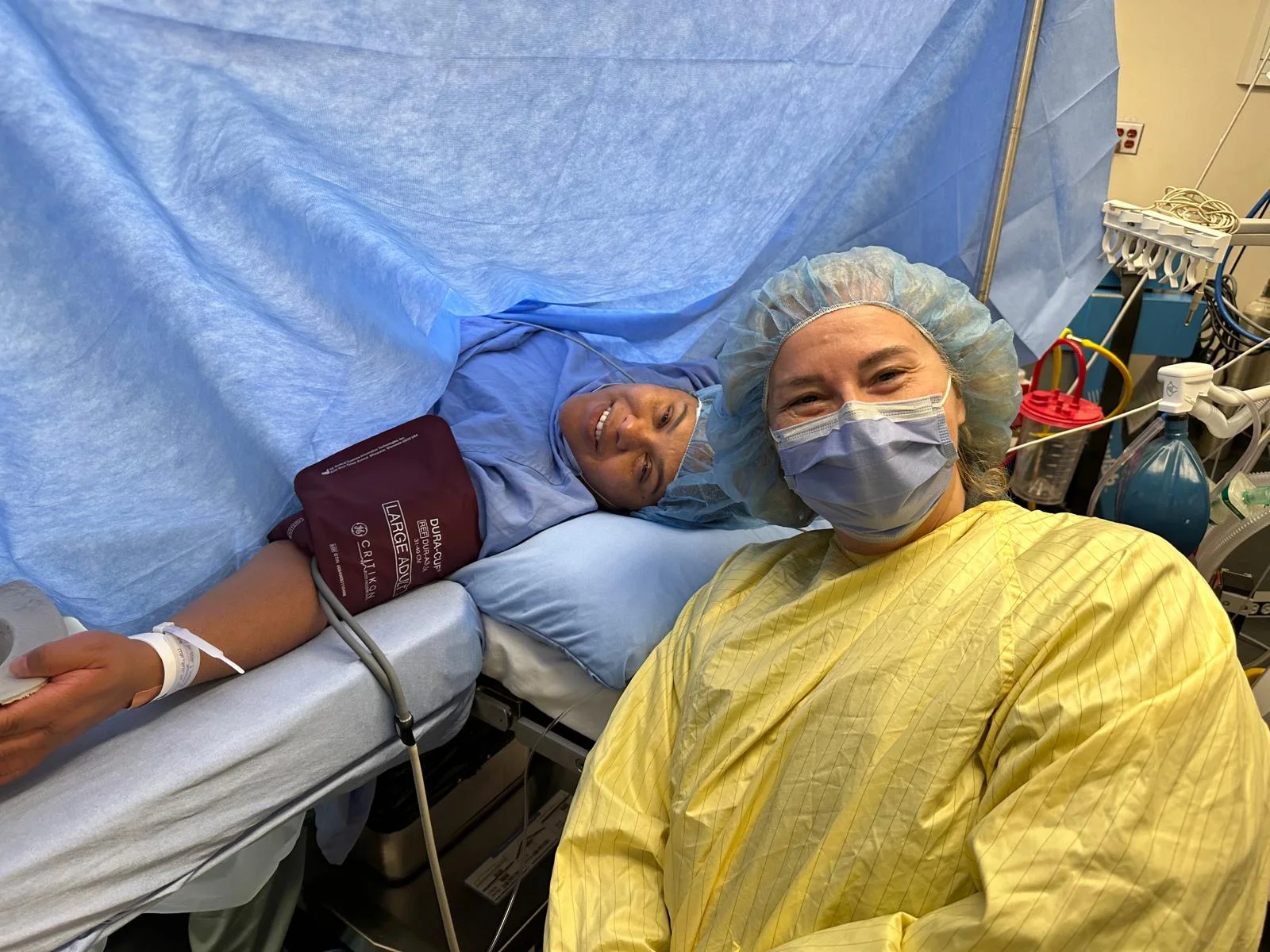 A women and her doula during a c-section. The new momma is lying down draped from her neck down giving birth via cesarean.